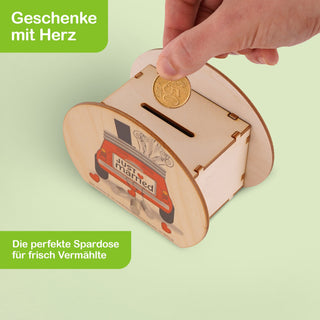 Rechteckige Spardose aus hellem Holz mit abgerundeten Seiten. Auf der Vorderseite ist ein Auto mit einem „Just Married“-Schild und zwei roten Herzen abgebildet. Eine Hand hält eine Münze über den Schlitz an der Oberseite der Spardose. Oben links steht in weißer Schrift auf grünem Hintergrund: „Geschenke mit Herz“. Unten links steht: „Die perfekte Spardose für frisch Vermählte“.