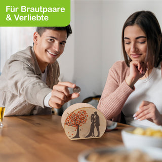 Ein Paar sitzt an einem Tisch. Der Mann hält eine Münze über eine Spardose mit Hochzeitsmotiv. Die Spardose zeigt einen Baum mit roten Blättern und die schwarze Silhouette eines Brautpaares. Oben links im Bild steht in weißer Schrift auf grünem Hintergrund: „Für Brautpaare & Verliebte“.
