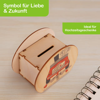 Rechteckige Spardose aus hellem Holz mit abgerundeten Seiten. Auf der Vorderseite ist ein rotes Auto mit einem „Just Married“-Schild abgebildet. Oben befindet sich ein Schlitz zum Einwerfen von Geld. Die Spardose steht auf einer hellen Holzoberfläche, im Hintergrund liegen ein Notizbuch und ein Stift. Im Bild steht: „Symbol für Liebe & Zukunft“ und „Ideal für Hochzeitsgeschenke“.
