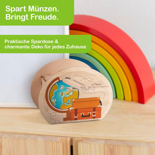 Hölzerne Spardose in Form eines Koffers mit einem aufgemalten Globus und Flugzeug. Im Hintergrund befindet sich ein halbkreisförmiges, buntes Regenbogen-Dekorationsstück. In der oberen linken Ecke steht in weißer Schrift auf grünem Hintergrund: „Spart Münzen. Bringt Freude.“ Darunter: „Praktische Spardose & charmante Deko für jedes Zuhause“.