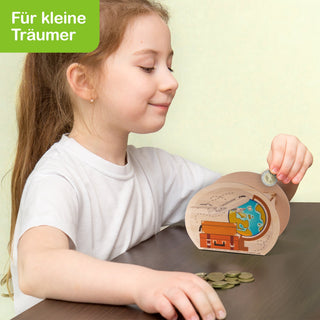 Ein Kind sitzt an einem Tisch und steckt eine Münze in eine Spardose mit Schatztruhen- und Weltkartenmotiv. Auf dem Tisch liegen weitere Münzen. In der oberen linken Ecke steht auf grünem Hintergrund der Text „Für kleine Träumer“.