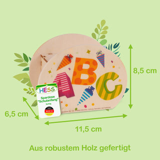 Spardose aus robustem Holz mit den Maßen 6,5 × 11,5 × 8,5 cm. Auf der Vorderseite sind die bunten Buchstaben A, B und C mit kleinen Schultüten verziert. Links unten befindet sich ein Etikett mit der Aufschrift: ‚HESS Spardose ‚Schulanfang‘ farbig – 100% MADE IN GERMANY‘. Im Hintergrund sind grüne Blätter als Dekoration zu sehen. Am unteren Bildrand steht: ‚Aus robustem Holz gefertigt‘.