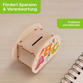 Kleine, rechteckige Holzspardose mit einem Schlitz oben zum Einwerfen von Münzen. Die Vorderseite ist bunt bemalt mit den Buchstaben A, B und C in verschiedenen Farben und Mustern. Links oben steht in einem grünen Feld: ‚Fördert Sparsinn & Verantwortung‘, rechts daneben: ‚Praktisch und dekorativ‘. Im Vordergrund unten rechts ist ein Spiralblock teilweise sichtbar.