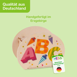 Runde, handgefertigte Spardose aus Holz mit bunten Buchstaben A, B und C sowie kleinen Schultüten als Dekor. Oben links im Bild steht in einem grünen Feld ‚Qualität aus Deutschland‘, darunter in grüner Schrift ‚Handgefertigt im Erzgebirge‘. Unten rechts ist ein Etikett mit der Aufschrift: ‚Hess – Spardose ‚Schulanfang‘ farbig – 100% Made in Germany‘.