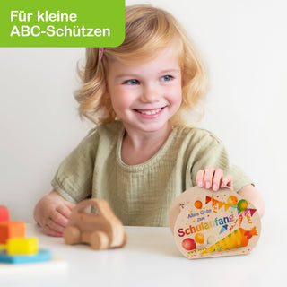 Ein kleines Kind sitzt an einem Tisch und hält ein buntes Buch mit der Aufschrift ‚Alles Gute zum Schulanfang‘. Auf dem Tisch liegen ein Holzspielzeugauto und mehrere bunte Bauklötze. Oben links im Bild steht in einem grünen Feld: ‚Für kleine ABC-Schützen‘.