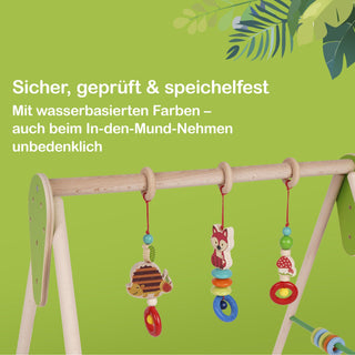 Holzspielbogen für Babys mit drei hängenden Spielzeugen. Der Hintergrund ist grün, oben rechts sind Blätter zu sehen. Ein Text auf dem Bild informiert: ‚Sicher, geprüft & speichelfest – Mit wasserbasierten Farben, auch beim In-den-Mund-Nehmen unbedenklich.‘