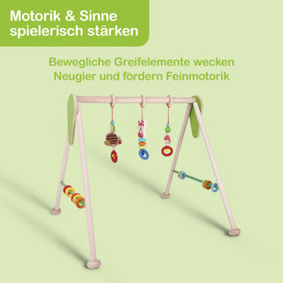 Hölzernes Babyspielgerät mit vier Beinen und einer oberen Querstange. Daran hängen bunte Greifelemente wie Ringe und Figuren in verschiedenen Farben und Formen. Der Hintergrund ist hellgrün. Oben links steht in einem grünen Feld: ‚Motorik & Sinne spielerisch stärken‘. Darunter in grüner Schrift: ‚Bewegliche Greifelemente wecken Neugier und fördern Feinmotorik‘.