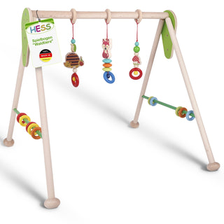 Hölzernes Spielbogen-Gestell mit vier Beinen und einer oberen Querstange. Daran hängen drei bunte Spielzeuge: ein Igel, eine Eule und ein Pilz – jeweils an bunten Ringen befestigt. An den Seiten befinden sich zusätzliche bunte Ringe zum Spielen. Ein Etikett am linken Bein trägt die Aufschrift ‚HESS Spielbogen ‚Waldtiere‘ 100% MADE IN GERMANY‘.“