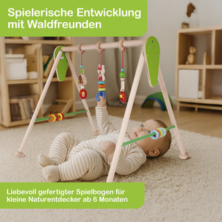 Ein Baby liegt auf dem Rücken auf einem Teppich und spielt mit einem hölzernen Spielbogen. Am Bogen hängen bunte Spielzeuge in verschiedenen Formen, die das Baby greifen kann. Im Hintergrund steht ein Regal mit Büchern und Gegenständen. Text im Bild: „Spielerische Entwicklung mit Waldfreunden“ und „Liebevoll gefertigter Spielbogen für kleine Naturentdecker ab 6 Monaten“.