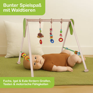 Ein Baby liegt auf dem Bauch auf einer grünen Matte und spielt mit einem hölzernen Spielgestell, an dem bunte Waldtier-Spielzeuge wie Fuchs, Igel und Eule hängen. Oben links im Bild steht: ‚Bunter Spielspaß mit Waldtieren‘. Unten steht: ‚Fuchs, Igel & Eule fördern Greifen, Tasten & motorische Fähigkeiten‘.