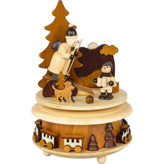 Holzspieluhr mit winterlicher Szene, die zwei Figuren zeigt – der Weihnachtsmann  auf Skiern und eine andere, die ein Geschenk hält. Ein Hund und mehrere Bäume sind ebenfalls zu sehen. Die Basis der Spieluhr ist mit kleinen Holzfiguren von Zügen und Bäumen verziert.
