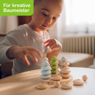 Ein Kind spielt mit einem Holzspielzeug, bei dem verschiedenfarbige und unterschiedlich große Ringe auf senkrechte Stäbe gesteckt werden. Im Hintergrund ist unscharf ein Zimmer mit Heizkörper und Fenster zu sehen. Oben links steht in weißer Schrift auf grünem Hintergrund: „Für kreative Baumeister“.