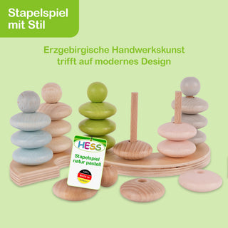 Ein Stapelspiel aus Holz in sanften Pastellfarben. Mehrere Holzstäbe tragen verschiedenfarbige Ringe, darunter Blau, Grün und Rosa. Oben links steht in einem grünen Feld: „Stapelspiel mit Stil“. Darunter: „Erzgebirgische Handwerkskunst trifft auf modernes Design“. In der Bildmitte ist ein Etikett mit der Aufschrift „HESS Stapelspiel natur pastell“ und dem Hinweis „100% Made in Germany“.