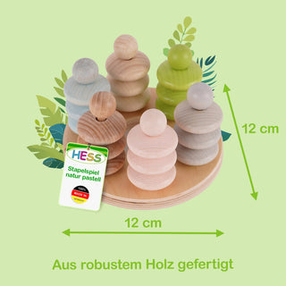 Ein Stapelspiel aus robustem Holz der Marke HESS in natürlichen Pastellfarben. Es steht auf einer runden Basis mit 12 cm Durchmesser und besteht aus mehreren stapelbaren Elementen in Blau, Grün und Naturholz. Die Gesamthöhe beträgt ebenfalls 12 cm. Ein Etikett zeigt die Aufschrift „HESS“, „Stapelspiel natur pastell“ und „Top Qualität Made in Germany“. Im Hintergrund sind stilisierte Blätter zu sehen.