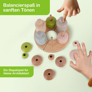Ein Stapelspiel aus Holz mit verschiedenfarbigen Ringen und senkrechten Stäben. Zwei Kinderhände sind sichtbar, eine greift nach einem der Ringe. Text im Bild: „Balancierspaß in sanften Tönen“ und „Ein Stapelspiel für kleine Architekten!“.