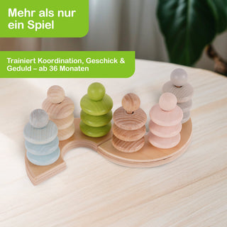 Ein Holzspielzeug mit mehreren stapelbaren Figuren in den Farben Blau, Grün, Naturholz und Rosa. Die Figuren stehen auf einer geschwungenen Holzplatte. Im Hintergrund ist eine Pflanze zu sehen. Text im Bild: „Mehr als nur ein Spiel – Trainiert Koordination, Geschick & Geduld – ab 36 Monaten“.