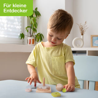 Ein kleines Kind mit gelbem T-Shirt sitzt an einem Tisch und spielt mit Holzspielzeug. Im Hintergrund sind eine Pflanze, ein Fenster mit Jalousien, eine weiße Vase und ein Bild an der Wand zu sehen. Oben links steht in weißer Schrift auf grünem Hintergrund: „Für kleine Entdecker“.