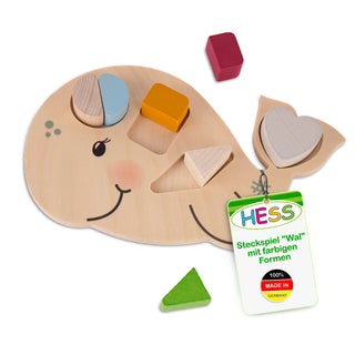Hölzernes Steckspiel in Form eines Wals mit farbigen geometrischen Formen. Der Wal hat Aussparungen für ein blaues Halbkreis, ein gelbes Quadrat, ein rotes Rechteck und ein grünes Dreieck. Einige Formen sind bereits eingesetzt. Am Spielzeug ist ein Etikett befestigt mit der Aufschrift: ‚HESS Steckspiel ‚Wal‘ mit farbigen Formen – 100% MADE IN GERMANY‘.