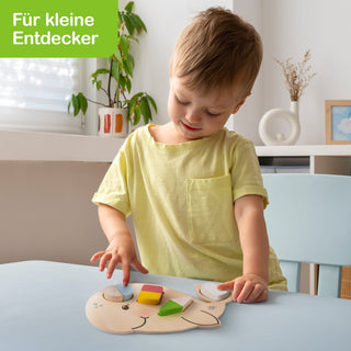 Ein kleines Kind sitzt an einem Tisch und spielt mit einem Holzpuzzle in Tierform. Das Puzzle besteht aus bunten Formen, die das Kind mit den Händen anordnet. Im Hintergrund sind eine Pflanze, ein weißer runder Dekorationsgegenstand und ein gerahmtes Bild zu sehen. Oben links steht auf grünem Hintergrund: ‚Für kleine Entdecker‘.