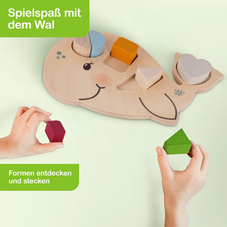 Holzspielzeug in Form eines Wals mit Aussparungen für farbige Formen: blaues Oval, orangefarbenes Quadrat, weißes Herz und beiges Dreieck. Zwei Hände halten jeweils eine rote und eine grüne Form. Oben links steht: ‚Spielspaß mit dem Wal‘, unten links: ‚Formen entdecken und stecken‘.