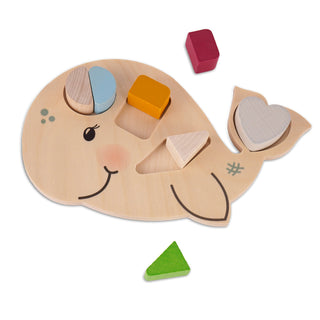 Hölzernes Steckpuzzle in Form eines lächelnden Wals mit rosa Wangen. Auf dem Wal sind verschiedene bunte Holzformen platziert: ein halbkreisförmiges blaues und holzfarbenes Stück, ein gelbes Quadrat, ein holzfarbenes Dreieck und ein weißes Herz. Neben dem Wal liegen ein rotes Quadrat und ein grünes Dreieck.