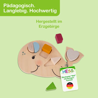 Holzspielzeug in Form eines Wals, gestaltet als Steckspiel mit farbigen Formen: grünes Dreieck, rotes Quadrat, gelbes Rechteck sowie zwei halbrunde Teile in Blau und Weiß. Die Formen passen in passende Aussparungen im Wal. Oben links steht auf grünem Hintergrund: ‚Pädagogisch. Langlebig. Hochwertig‘. In der Mitte: ‚Hergestellt im Erzgebirge‘. Unten rechts befindet sich ein Etikett mit dem Logo ‚HESS‘ und dem Text: ‚Steckspiel ‚Wal‘ mit farbigen Formen – 100% Made in Germany‘.