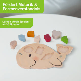 Holzpuzzle in Form eines Wals mit bunten geometrischen Formen: blauer Halbkreis, orangefarbenes Quadrat, pinkes Dreieck, rotes Rechteck, lilafarbenes Herz und grünes Dreieck. Der Text auf dem Bild lautet: ‚Fördert Motorik & Formenverständnis‘ sowie ‚Lernen durch Spielen – ab 36 Monaten‘.