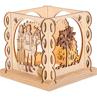 Der kleine hölzerne Teelichthalter in Natur ist quadratisch und mit mehreren Rundungen an den Außenseiten und mit verschiedenen Blenden versehen. Zu sehen ist hier das Zauberbäumchen und das Prinzenpaar mit Pferd. Dieses Bild zeigt den beleuchteten Teelichthalter. Bild 2.