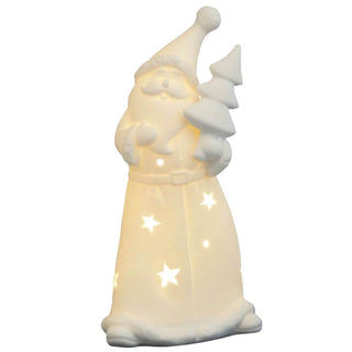 Ein weißer Weihnachtsmann aus Porzellan ist beleuchtet und hält einen stilisierten Weihnachtsbaum in der linken Hand. Sein Mantel ist mit sternförmigen Aussparungen versehen, durch die warmes Licht scheint.