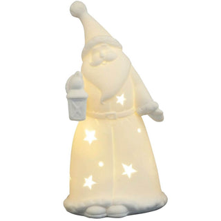 Ein weißer Weihnachtsmann aus Porzellan trägt einen langen Mantel und einen spitzen Hut. Er hält eine Laterne in der linken Hand. Sternförmige Aussparungen im Mantel lassen warmes Licht hindurchscheinen, wodurch die Figur von innen beleuchtet wirkt.