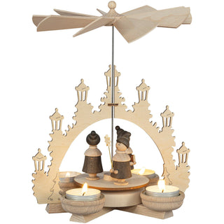 Holzpyramide mit vier Teelichthaltern, verziert mit geschnitzten Figuren von Kurrendesängern unter einem Bogen aus Laternen. Oben befindet sich ein Propeller, der durch die Wärme der Kerzen angetrieben wird.