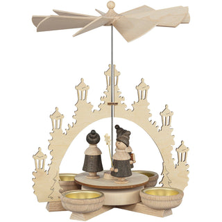 Holzpyramide mit vier Teelichthaltern, verziert mit geschnitzten Figuren von Kurrendesängern unter einem Bogen aus Laternen. Oben befindet sich ein Propeller, der durch die Wärme der Kerzen angetrieben wird.