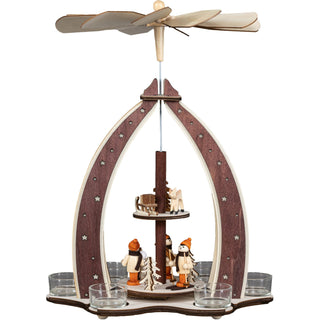 Ein Weihnachtspyramiden-Dekorationsstück aus Holz mit einem drehbaren Flügelrad oben, das durch die Wärme von Teelichten angetrieben wird. Die Pyramide zeigt Winterkinder, Reh, Schlitten und Bäumen auf zwei Ebenen, umgeben von sternverzierten Bögen.