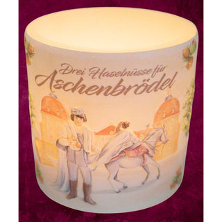 Ein Windlicht aus Wachs mit einem Durchmesser von 10 cm. Auf dem Windlicht ist eine Szene aus dem Märchen „Drei Haselnüsse für Aschenbrödel®“ abgebildet: Der Prinz in mittelalterlicher Kleidung blickt auf eine Prinzessin, die auf einem weißen Pferd sitzt. Im Hintergrund ist das Schloß zu sehen. Oben auf dem Windlicht steht in geschwungener Schrift „Drei Haselnüsse für Aschenbrödel“. beleuchtet