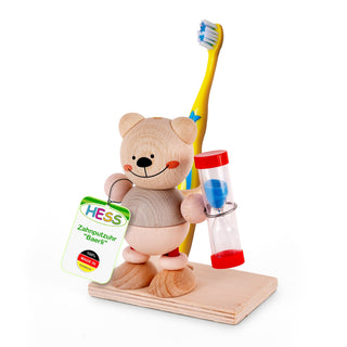 Eine hölzerne Bärenfigur steht auf einer rechteckigen Holzplatte. Der Bär hält in der rechten Hand eine Sanduhr mit roten Enden und blauem Sand, in der linken ein Schild mit der Aufschrift „HESS Zahnputzuhr ‚Baerli‘ 100% MADE IN GERMANY“. Hinter dem Bären steht eine gelbe Zahnbürste mit blauen Sternen und weißen Borsten.