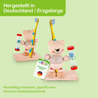 Zwei Holzfiguren in Bärenform stehen auf einer rechteckigen Holzplatte. Jede Figur hält eine Zahnbürste und eine Sanduhr. Eine Figur trägt ein Etikett mit der Aufschrift „HESS Zahnpfutzuh ‚Bärin‘“. Oben links im Bild steht „Hergestellt in Deutschland / Erzgebirge“ auf grünem Hintergrund, unten links „Nachhaltig produziert, geprüft nach höchsten Qualitätsstandards“.