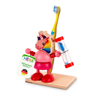 Eine bunte Einhornfigur aus Holz mit rosa Körper und regenbogenfarbenem Kopf steht auf einer Holzplatte. In einer Hand hält sie eine kleine Sanduhr mit blauem Sand und roten Enden, in der anderen ein Schild mit der Aufschrift „HESS Zahnputzuhr ‚Einhorn‘ 100% MADE IN GERMANY“. Hinter der Figur befindet sich eine gelbe Zahnbürste mit blauen Borsten.