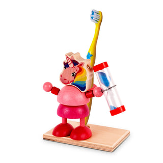 Ein bunter Zahnbürstenhalter aus Holz in Form eines Einhorns mit Regenbogenmuster auf dem Kopf. Das Einhorn steht auf einer Holzplatte und hält eine Sanduhr mit blauem Sand in der rechten Hand. Hinter dem Einhorn ist eine gelbe Zahnbürste mit blauen Borsten befestigt.
