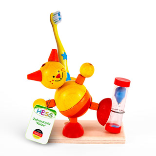 Eine bunte Holzfigur in Form einer Katze balanciert auf einem Bein. Die Katze hält eine Zahnbürste in der einen Hand und ein Schild in der anderen, auf dem steht: „HESS Zahnputzuhr ‚Katze‘ 100 % MADE IN GERMANY“. Neben der Figur steht eine kleine Sanduhr mit blauem Sand.