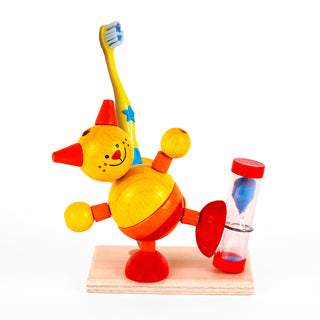 Ein buntes Holzspielzeug in Form einer Katze. Die Katze ist gelb bemalt und hat rote Ohren, Füße und eine rote Nase. Auf ihrem Rücken ist eine gelbe Zahnbürste befestigt. Sie steht auf einem Bein auf einer Holzplatte. Daneben befindet sich eine kleine Sanduhr mit blauem Sand und roten Endkappen.