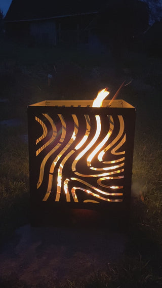 Das Bild zeigt eine Metall-Feuerschale mit einem dekorativen Ausschnittmuster, aus der Flammen emporsteigen. Die Feuerschale steht im Freien auf einer Grasfläche, und es ist Nacht.