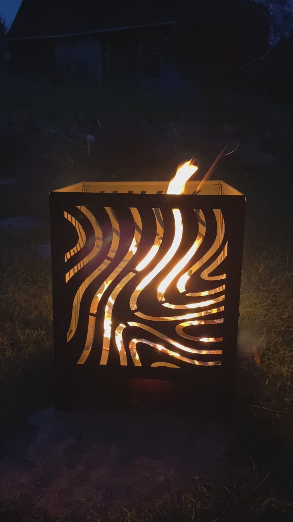 Das Bild zeigt eine Metall-Feuerschale mit einem dekorativen Ausschnittmuster, aus der Flammen emporsteigen. Die Feuerschale steht im Freien auf einer Grasfläche, und es ist Nacht.