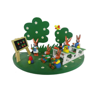 Auf einem grünfarbenen Sockel mit Blumen befindet sich eine Hasenschule mit farbigen Osterhasen und einer Schultafel. Im Hintergrund befinden sich zwei Bäumchen.