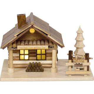 Kleines Räucherhaus aus Holz mit dem Schild „Gasthof zur wilden Sau“. Das Haus hat ein schräges Dach mit Schornstein, zwei Fenster und einen Holzstapel davor. Rechts daneben befindet ein geschnitzter Baum und ein Spanferkel hängt am Spieß.
