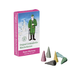 Verpackung der Original Crottendorfer Räucherkerzen ‚Bunte Mischung‘, gestaltet in Blau und Pink. Auf der Vorderseite ist eine winterliche Szene mit einem Mann in grüner Kleidung und Spazierstock vor einem Kirchturm abgebildet. Darunter steht ‚Original Crottendorfer Räucherkerzen‘ sowie ‚Bunte Mischung – Variety Pack – Pot - pourri‘. Neben der Verpackung liegen sechs verschiedenfarbige, konische Räucherkerzen: rot, grau, gelb, grün, blau und ein dunkleres grün.