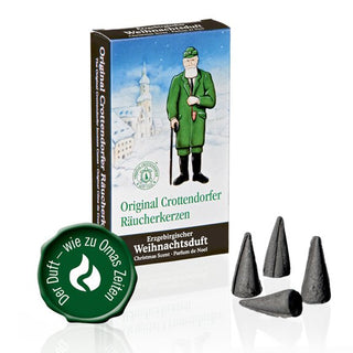 Verpackung der Original Crottendorfer Räucherkerzen ‚Erzgebirgischer Weihnachtsduft‘, 24 Stück. Die Schachtel zeigt eine winterliche Szene mit einem Mann in grüner Kleidung und Spazierstock vor einem verschneiten Dorf. Aufdruck mit Produktnamen in Deutsch, Englisch und Französisch. Links ein grünes Siegel mit ‚Der Duft wie zu Omas Zeiten‘. Davor vier schwarze, konische Räucherkerzen.