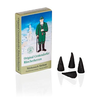 Verpackung der Original Crottendorfer Räucherkerzen ‚Weihrauch Melisse‘ in hellen Farben mit winterlicher Illustration: Ein Mann in traditioneller Kleidung steht vor einer verschneiten Dorfkulisse. Auf der Vorderseite der Schachtel steht ‚Original Crottendorfer Räucherkerzen‘ und darunter ‚Weihrauch Melisse‘. Vor der Verpackung liegen vier schwarze, konisch geformte Räucherkerzen.