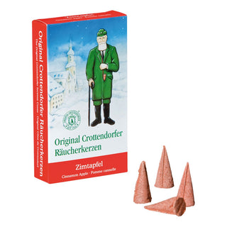 Verpackung der Original Crottendorfer Räucherkerzen ‚Zimtapfel‘ mit winterlicher Illustration eines verschneiten Dorfs und einer Figur in grüner Kleidung. Die Schachtel ist blau-rot gestaltet, trägt den Produktnamen sowie die Übersetzungen ‚Cinnamon Apple‘ und ‚Pomme cannelle‘. Vor der Verpackung liegen vier rötlich-braune, konische Räucherkerzen.