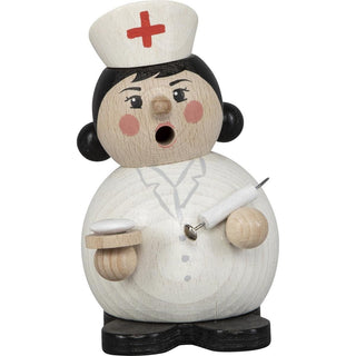 Räuchermännchen „Krankenschwester“ zeigt eine Holzfigur mit rundem Körper, bemalt als Krankenschwester mit weißer Haube und rotem Kreuz. Die Figur hat ein offenes Mundloch für den Rauch der Räucherkerze, hält eine Spritze in der einen Hand und ein Tablett in der anderen.