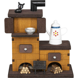 Der Räucherofen aus Massivholz besteht aus 2 Ebenen. Auf der oberen Ebene sitzt eine kleine, schwarze Katze. Auf der unteren Ebene steht eine Kaffeekanne. Vor dem Ofen liegen 2 Holzstapel und in der Mitte, vor dem Ofen steht noch eine kleines Bänkchen_1